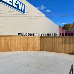 7 Below Custom Outdoor Signs that read 'Welcome to Franklin Square'. Sign is on top of a light brown wooden fence with a concrete wall behind.
