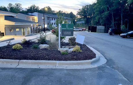 Landscape in front of an apartment complex with a post and panel sign. Green background with white letters reading, "Emery Village, Leasing Center".