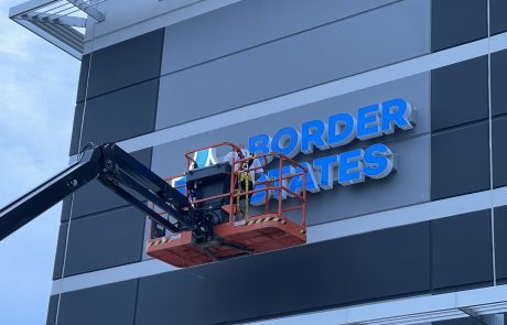 Outdoor Sign being installed by a worker on a black and red crane. black and grey outdoor wall with blue letters reading Border States.